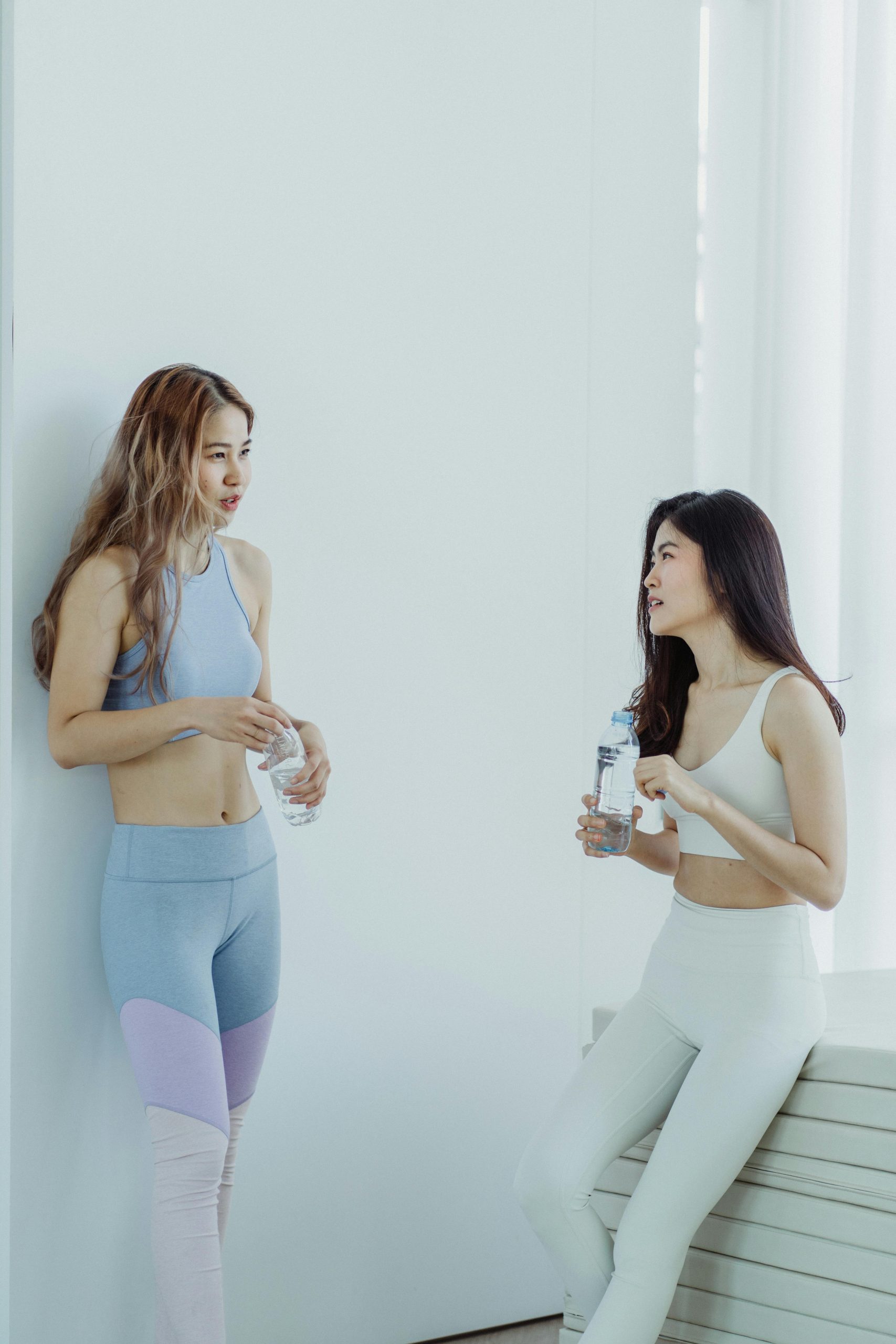 Two Asian women in yogawear talking and holding water bottles indoors.